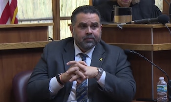 Un hombre con traje gris, corbata y barba permanece sentado con las manos entrelazadas en un estrado judicial