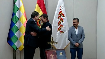 Dos hombres se abrazan frente a banderas, incluyendo la wiphala. Un tercer hombre sonríe a la derecha. Hay documentos y una cruz dorada en una mesa al frente