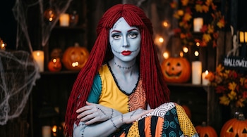 Mujer con piel azulada, costuras dibujadas, pelo rojo de estambre y vestido estampado, rodeada de calabazas y velas.
