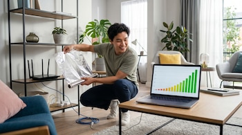Hombre asiático sonriente coloca papel aluminio detrás de un módem WiFi en una estantería. Una laptop muestra un gráfico de barras verdes indicando mejora de señal.