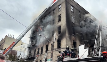 Edificio de ladrillo de varias plantas en llamas, con humo negro saliendo de las ventanas. Dos escaleras de camiones de bomberos y varios bomberos en acción