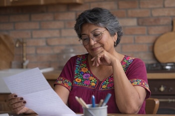 Mujer con cabello gris y lentes lee documento blanco. Viste blusa bordada. En mesa de madera, frente a pared de ladrillo y utensilios de cocina.