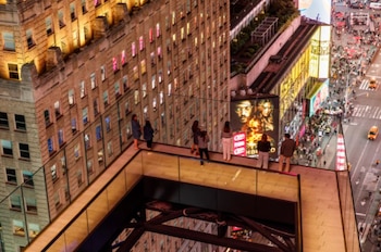 Vista aérea de personas en una terraza con barandillas de cristal observando Times Square por la noche, con edificios iluminados y calles concurridas