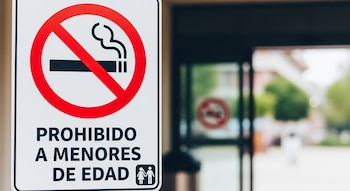 Un cartel blanco con borde negro en primer plano que muestra el símbolo universal de no fumar y el texto "PROHIBIDO A MENORES DE EDAD" con un icono de niños.