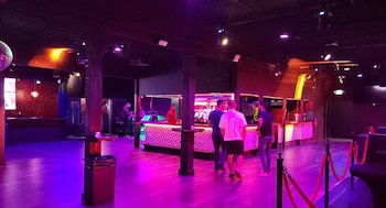 Interior de un club nocturno con luces moradas, personas junto a la barra, mesas altas y cuerdas de seguridad