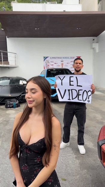 Una mujer con cabello largo y un hombre posan. Él sujeta un cartel con "Y EL VIDEO?". Detrás, dos coches deportivos y un letrero de "ELIMINATORIAS MUNDIAL 2026"