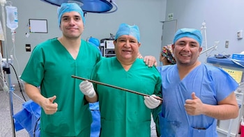 Tres cirujanos posan en un quirófano; el del centro, con uniforme verde y guantes, sujeta una varilla metálica. Los otros dos hacen la señal de aprobación