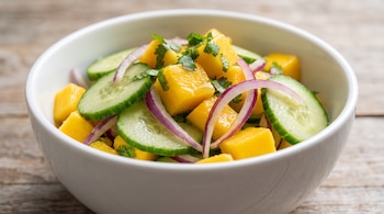 Plato blanco hondo con ensalada de cubos de mango, rodajas de pepino, plumas de cebolla morada y hojas de cilantro picado sobre una mesa de madera.