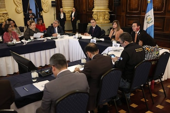 Un grupo de funcionarios sentados alrededor de mesas en forma de U en una sala con la bandera de Guatemala