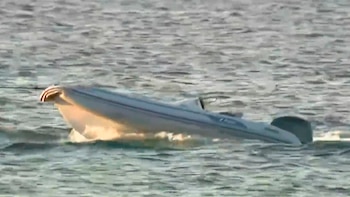 Un bote a motor blanco y gris oscuro se desplaza sobre el agua en un cuerpo de agua amplio. Se observan pequeñas ondas en la superficie del agua