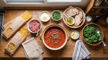 Mesa de madera con paquetes de pasta, olla de salsa de tomate, carne picada, queso rallado, rodajas de pan y hojas verdes en recipientes.