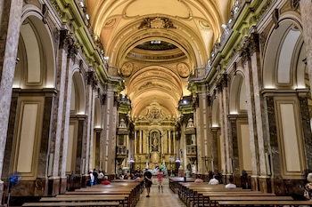 Interior de una gran iglesia con techo abovedado, columnas de mármol y un altar dorado al fondo. Varias personas observan el espacio y se sientan en los bancos