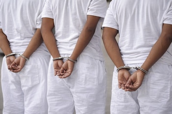 Tres personas de piel morena vistas de espaldas, vestidas con camisetas y pantalones cortos blancos, con las manos esposadas detrás de la espalda.