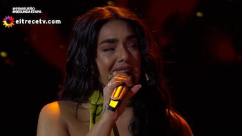 Primer plano de una mujer con cabello oscuro y un top verde, cantando con ojos cerrados en un micrófono dorado. Su rostro brilla bajo la luz cálida del escenario