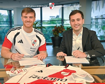 Dos hombres firman documentos sentados frente a una mesa de vidrio. En primer plano, una camiseta de fútbol blanca de River Plate con franja roja