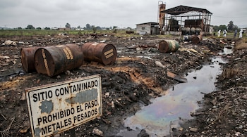 Sitio industrial contaminado con barriles de residuos tóxicos, un arroyo iridiscente y un letrero. Edificios industriales y personal con trajes protectores en el fondo.