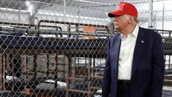 Donald Trump, con traje oscuro, camisa blanca y gorra roja, mira a la derecha frente a una valla metálica. Detrás, se ven literas a través de la cerca