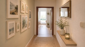 Pasillo interior con paredes claras, galería de cuadros de paisajes y botánica, alfombra beige, estante de madera, espejo, jarrón con ramas y vela encendida.