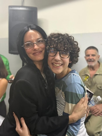 Oriana Sabatini con anteojos abraza a un joven de cabello rizado con anteojos. Un hombre mayor sonríe de fondo. Hay un parlante y una botella