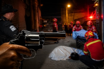 Mano que empuña un revólver en primer plano frente a una calle con socorristas, un policía y una persona tendida bajo una sábana.