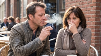 Un hombre sostiene un cigarrillo encendido con humo junto a una mujer que se tapa la nariz y la boca en una mesa exterior.