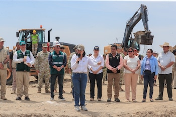 Una mujer habla por micrófono ante un grupo de personas con chalecos y uniformes militares; excavadora y bulldozer en terreno seco bajo cielo claro