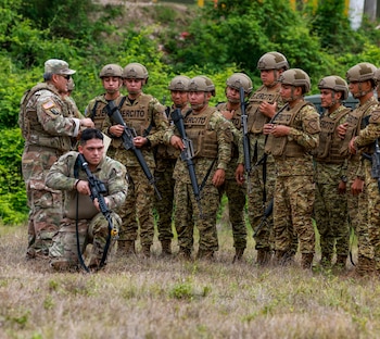 Un oficial militar instruye a soldados uniformados con rifles y chalecos tácticos en un campo. Un soldado se arrodilla en primer plano, con vegetación de fondo