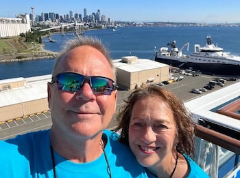 Selfie de un hombre con gafas de sol y una mujer sonriendo, con el horizonte de Seattle, su bahía y barcos en un día soleado de fondo
