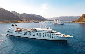 Un crucero blanco grande de Windstar Cruises se desplaza en agua azul clara. Al fondo, otro crucero de vela blanco y montañas con un pueblo costero