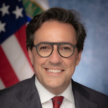 Hombre con gafas de montura oscura, traje gris, camisa blanca y corbata roja sonríe frente a la bandera de Estados Unidos
