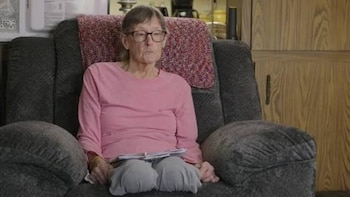 Una mujer con gafas y camiseta rosa que presenta amputación en ambas piernas permanece sentada en un sillón gris con papeles en el regazo