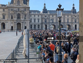 Una multitud de personas espera detrás de vallas metálicas en una plaza de piedra frente a la fachada clásica de un palacio con farolas