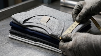 Manos con guantes industriales sostienen una cinta métrica sobre camisetas dobladas de color gris, azul marino y blanco en una mesa de metal.