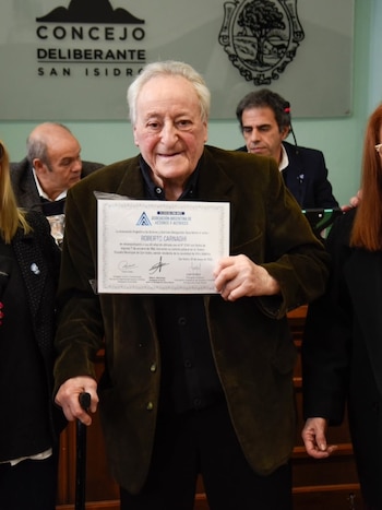 Hombre mayor de cabello blanco sonríe, sosteniendo un certificado y un bastón, frente a un letrero del Concejo Deliberante de San Isidro