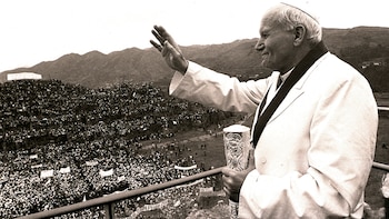 Un hombre, el papa Juan Pablo II, saluda con su mano derecha y sostiene un báculo con la izquierda, sobre una plataforma, ante una gran multitud y montañas