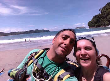Un hombre y una mujer sonriendo en un selfie, con una playa de arena, el mar con olas, dos personas en el agua y varias islas en el fondo