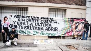 Tres mujeres sostienen en la calle un cartel de tela con un texto escrito y la ilustración del rostro de un hombre