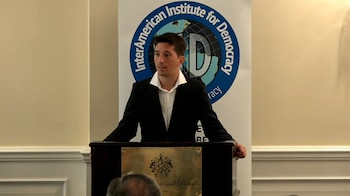 Hombre de traje negro y camisa blanca detrás de un podio de madera con placa dorada. Detrás, un banner con el logo del InterAmerican Institute for Democracy