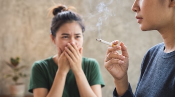 Una mano sostiene un cigarrillo encendido con humo y, al fondo, una mujer se cubre el rostro con las dos manos.
