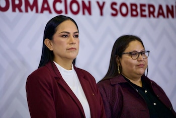 Dos mujeres de pie, una con blazer rojo y camisa blanca, otra con gafas y chaqueta estampada, delante de un muro blanco con texto rojo borroso