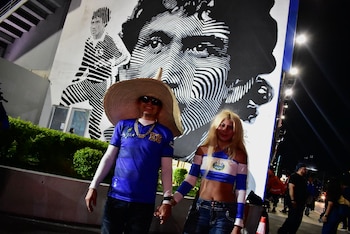 Un hombre con sombrero grande de paja y una mujer se toman de la mano frente a un mural en blanco y negro del rostro de un futbolista