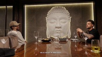 Dos hombres están sentados a los extremos de una mesa de madera con copas y vajilla frente a la escultura de una cabeza de Buda