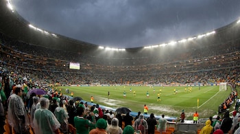 Vista panorámica de un estadio de fútbol con un partido en curso bajo lluvia intensa. Jugadores en el campo, gradas llenas de aficionados con ponchos y paraguas.