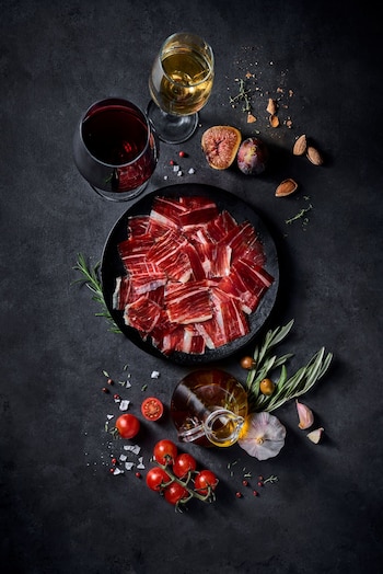 Plato negro con lonchas de jamón, copas de vino tinto y blanco, tomates, higos, ajo y aceitera en un fondo oscuro visto desde arriba
