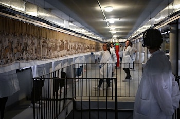 Extenso tapiz histórico en un pasillo iluminado. Varias personas con batas blancas inspeccionan el tapiz. Barandilla metálica negra
