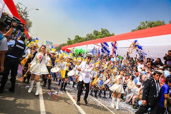 Desfile con banda escolar, director, estudiantes en uniformes con tambores, público con globos, y confeti cayendo en una calle