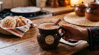 Jarro de barro con champurrado de chocolate y canela, sostenido por una mano, junto a un canasto con piezas de pan dulce sobre una mesa.
