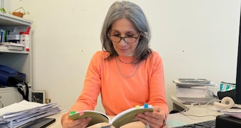 Mujer con cabello gris y gafas lee un libro con separadores de colores en un escritorio lleno de papeles, monitor y teclado