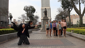 Nueve bailarines realizan una coreografía en un parque urbano. Dos interactúan en primer plano, mientras siete se alinean al fondo, frente a un monumento y árboles