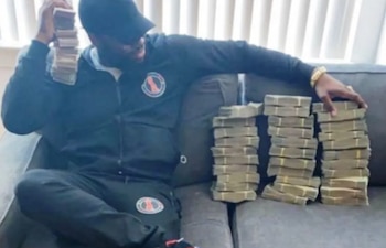 Hombre sentado en un sofá negro, viste gorra y sudadera oscuras. Sostiene un gran fajo de billetes en alto; tres grandes fajos de billetes sobre el sofá